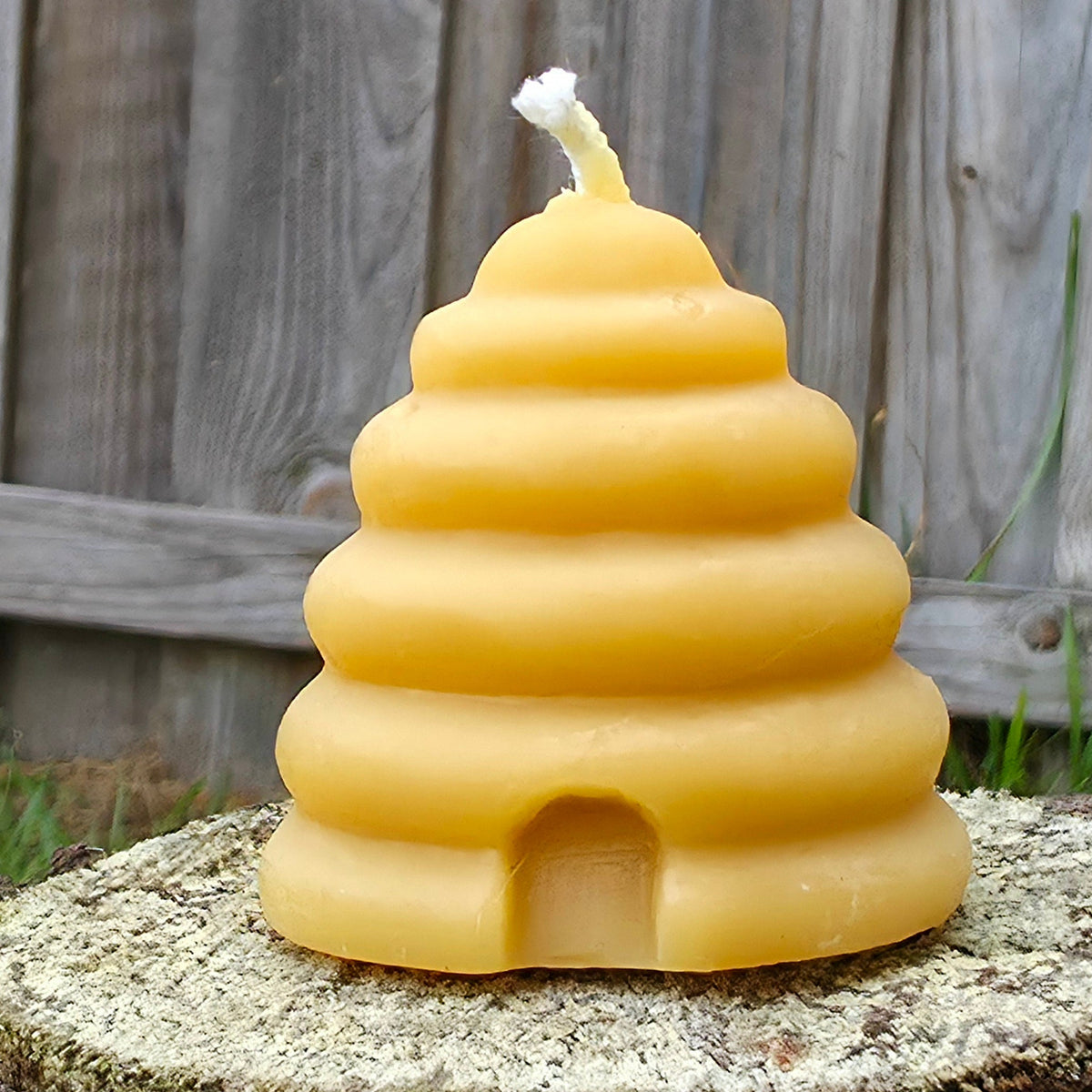 A yellow honeycomb-style beehive candle with a white wick, displayed on a tree stump with a wooden fence in the background.