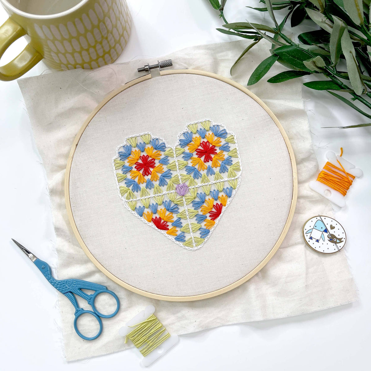 A heart-shaped embroidery design in a wooden hoop, surrounded by crafting tools including blue scissors, spools of thread, a small compass, and a mug on a white fabric background.