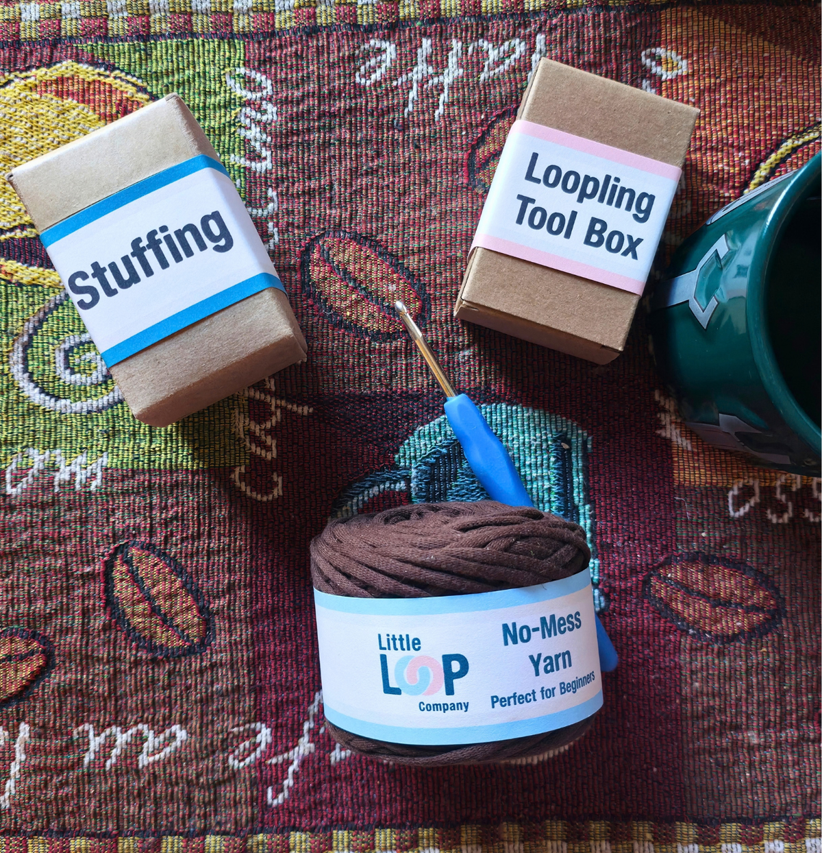 Beginner crochet kit flat lay with no-mess yarn, looping tool box, and stuffing on a coffee-themed tapestry.